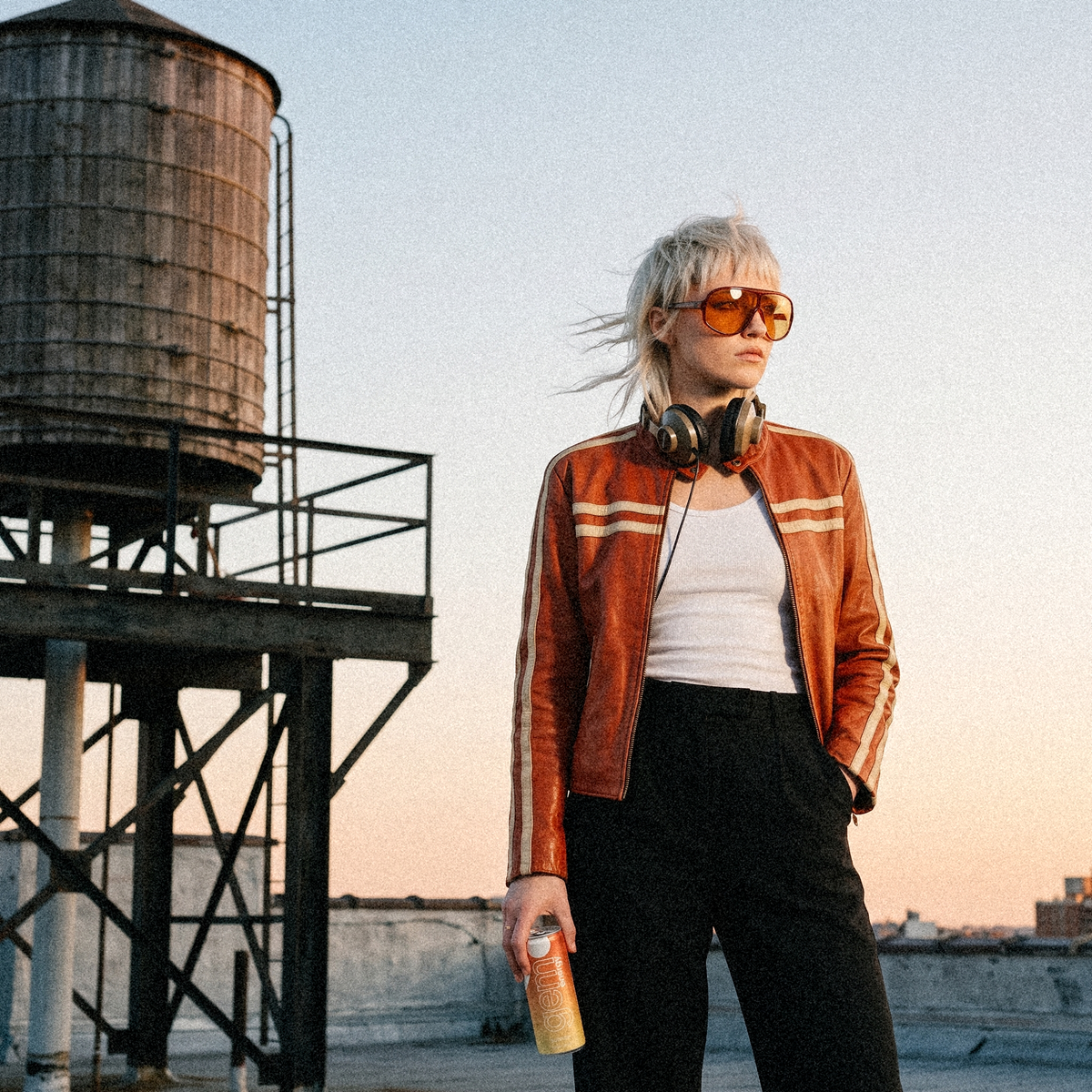 Young woman with a platinum mullet, orange shield sunglasses and a vintage leather racing jacket on a rooftop by a water tower, a can of Gem Energy Sunrise at her hip